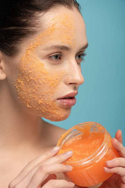 привлекательная женщина применяет желтый сахарный скраб из пластикового контейнера, изолированного на синий
