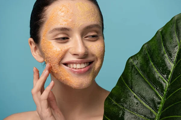 smiling girl with sugar scrub on face holding leaf, isolated on blue