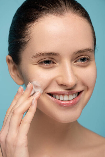 smiling young woman applying cleansing foam on face, isolated on blue