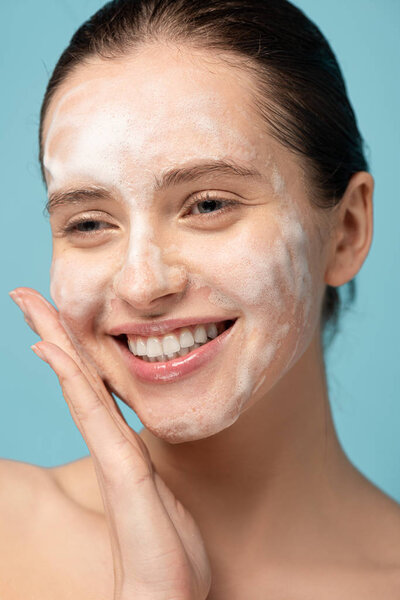 attractive happy girl applying cleansing foam on face, isolated on blue