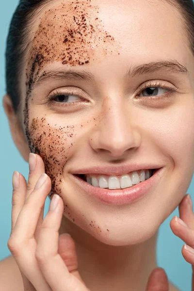 positive woman applying coffee scrub on face, isolated on blue