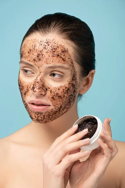 beautiful girl applying coffee scrub from plastic container, isolated on blue
