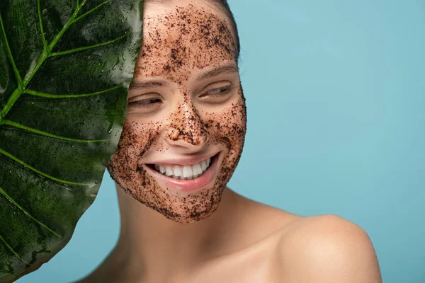 smiling young woman with coffee scrub on face, isolated on blue with leaf