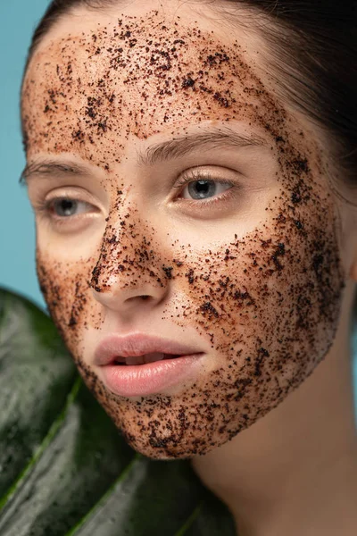 attractive girl with coffee scrub on face, isolated on blue with leaf