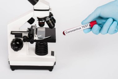 Beyaz üzerinde izole edilmiş mikroskop yakınında Coronavirus test harfleriyle numune tutan bilim adamının kırpılmış görüntüsü 