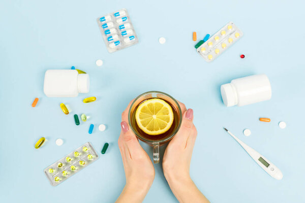 top view of woman holding cup of tea with sliced lemon near pills and digital thermometer on blue 