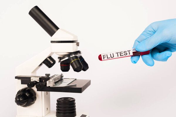 cropped view of scientist in latex glove holding test tube with flu test lettering near microscope isolated on white 