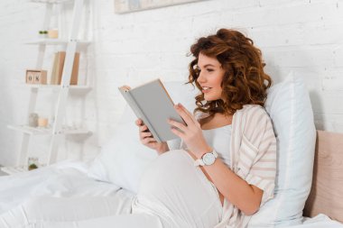 Yatakta kitap okurken gülümseyen çekici hamile kadın. 