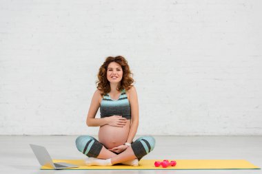Hamile kadın dizüstü bilgisayarın yanında karnına dokunurken kameraya gülümsüyor ve spor paspasındaki dambıllar...