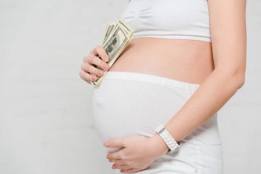 Cropped view of pregnant woman holding dollar banknotes and touching belly on grey background, concept of surrogacy maternity clipart