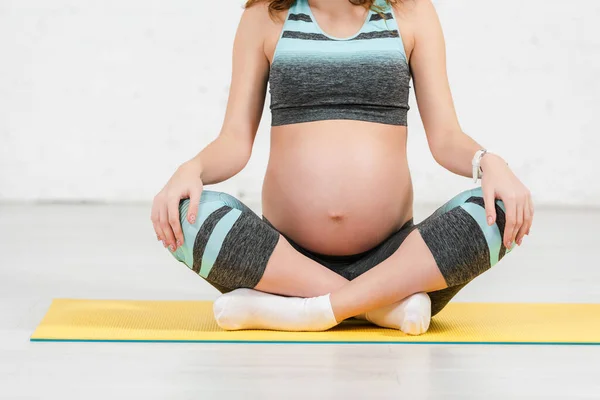 床の上のフィットネスマットに座っているスポーツウェアで妊娠中の女性の作物ビュー — ストック写真