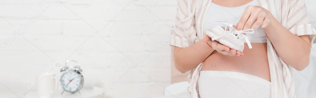 Cropped view of pregnant girl holding baby booties in bedroom, panoramic shot