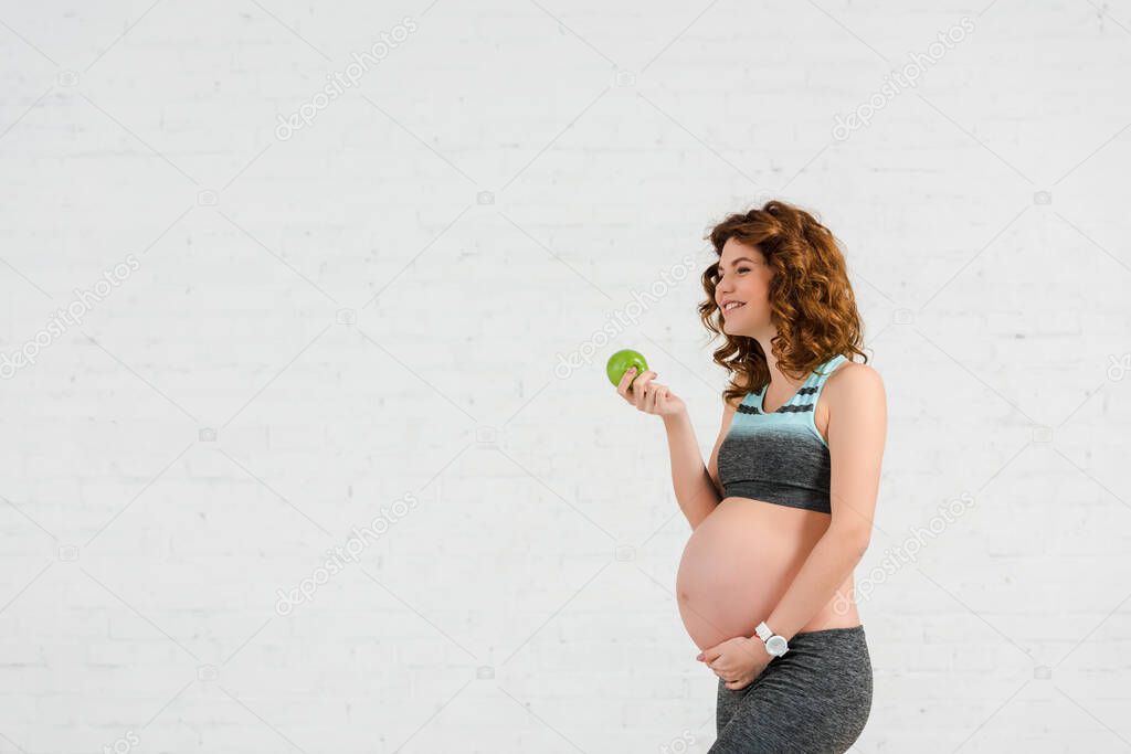 Side view of smiling pregnant woman holding apple on white background