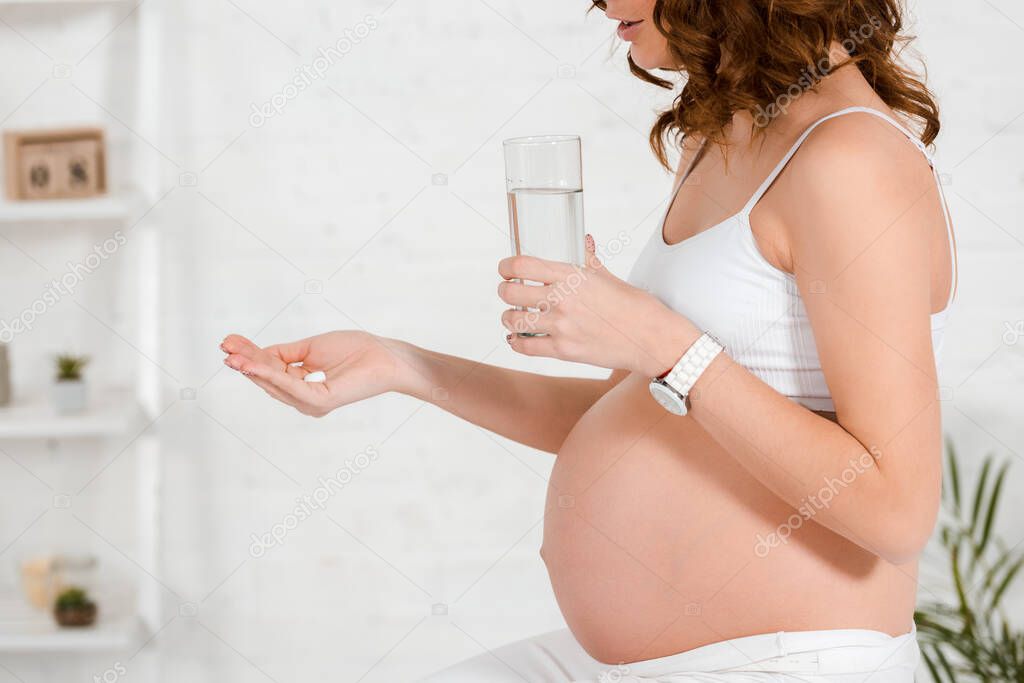 Cropped view of pregnant woman holding pill and glass of water in living room
