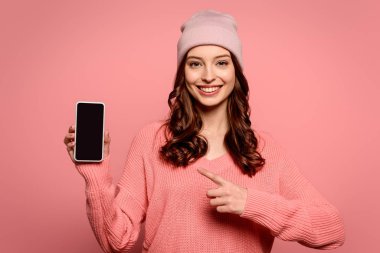 Mutlu kız pembe ekranda izole edilmiş kameraya bakarken boş ekranla akıllı telefonu işaret ediyor.
