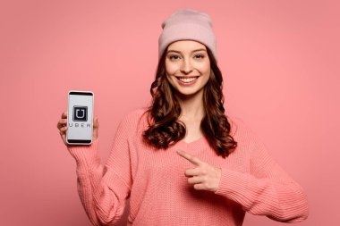 KYIV, UKRAINE - NOVEMBER 29, 2019: smiling girl pointing with finger at smartphone with Uber app on screen isolated on pink