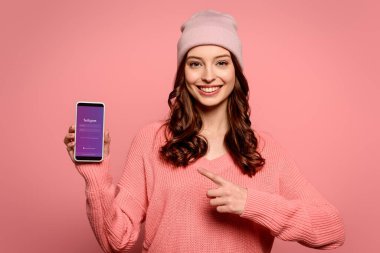 KYIV, UKRAINE - NOVEMBER 29, 2019: smiling girl pointing with finger at smartphone with Instagram app on screen isolated on pink
