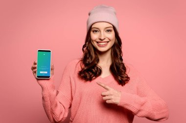KYIV, UKRAINE - NOVEMBER 29, 2019: happy girl pointing with finger at smartphone with Twitter app on screen isolated on pink