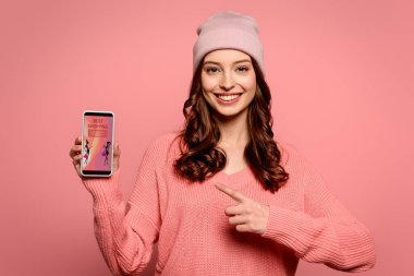 happy girl pointing with finger at smartphone with best shopping app on screen isolated on pink