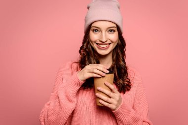 Neşeli kız pembe üzerinde izole olmak için elinde kahve tutarken kameraya gülümsüyor.