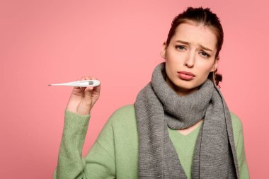 sick, displeased girl in warm showing thermometer isolated on pink clipart