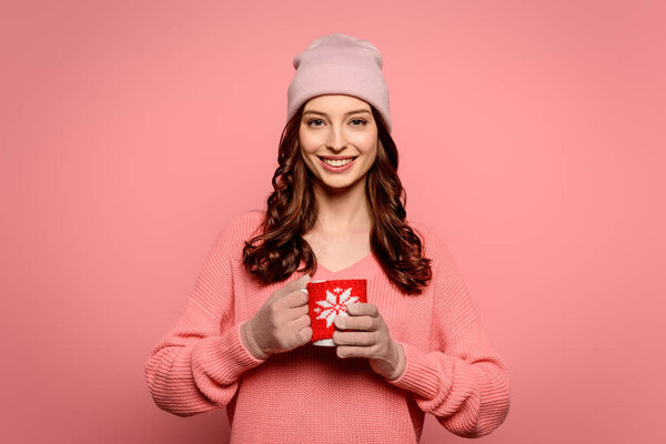 smiling girl in hat and gloves holding mug with warming drink isolated on pink