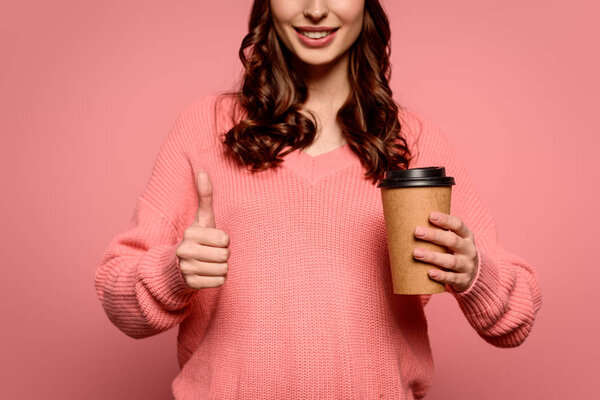 cropped view of smiling girl holding coffee to go and showing thumb up on pink background