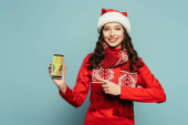 glückliches Mädchen in Weihnachtsmütze und rotem Pullover zeigt mit dem Finger auf Smartphone mit bester Shopping-App auf blauem Hintergrund
