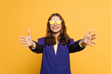 cheerful, stylish girl in yellow glasses showing welcome gesture isolated on yellow clipart