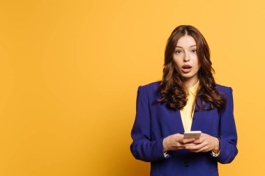 Sarı arka planda akıllı telefonu tutarken kameraya bakan şok kız.