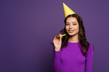 Mor arka plandaki kameraya bakarken elinde parti şapkası olan gülümseyen kız.