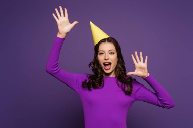 excited girl in party cap, with open mouth, gesturing with hands on purple background clipart