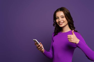 Akıllı telefonu tutan ve mor arka planda baş parmağını gösteren neşeli kız.