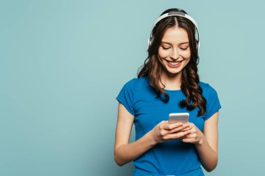 Kablosuz kulaklıklı neşeli kız mavi arka planda boş ekranlı akıllı telefonu tutarken başparmağını kaldırıyor.