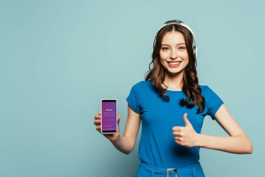 Kyiv, Ukrayna - 29 Kasım 2019: Mavi arka planda Instagram uygulaması ile akıllı telefonu tutarken parmaklarını gösteren kablosuz kulaklıklı mutlu kız