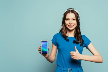 Kablosuz kulaklıklı mutlu kız mavi arkaplandaki ekranda uygulamalı akıllı telefonu tutarken başparmağını kaldırıyor.
