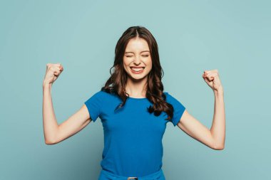 excited girl showing winner gesture with closed eyes while smiling on blue background clipart