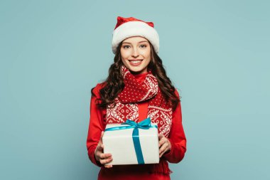 Noel Baba şapkalı mutlu kız mavi kameraya bakarken hediye kutusunu gösteriyor.