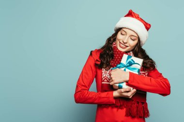Noel Baba şapkalı mutlu kız mavi arka planda kapalı gözleri olan hediye kutusuna sarılıyor.