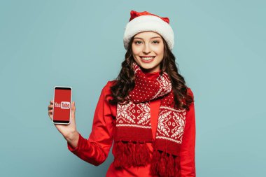 Kyiv, Ukrayna - 29 Kasım 2019: Noel Baba şapkalı ve kırmızı kazaklı mutlu kız Youtube uygulamalı akıllı telefonu mavi arka planda gösteriyor