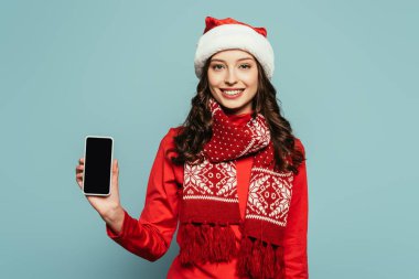 Noel Baba şapkalı ve kırmızı kazaklı gülümseyen kız mavi arka planda boş ekranlı akıllı telefon gösteriyor.