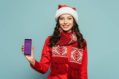 Kyiv, Ukrayna - 29 Kasım 2019: Noel Baba şapkalı ve kırmızı kazaklı neşeli kız Instagram uygulamalı akıllı telefonu mavi arka planda gösteriyor