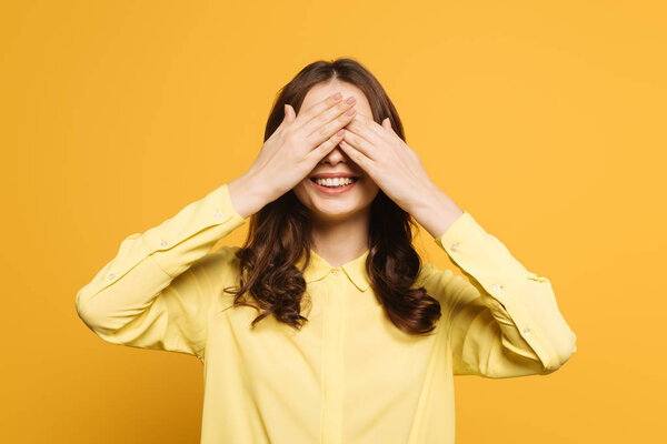 cheerful girl covering eyes with hand on yellow background