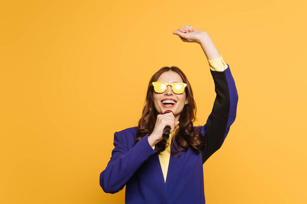 young singer in yellow glasses singing with raised hand isolated on yellow