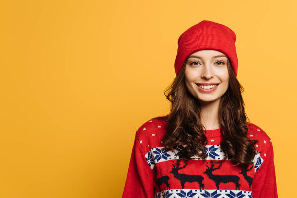 happy girl in hat and red ornamental sweater smiling at camera isolated on yellow