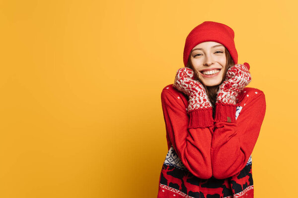 happy girl in red ornamental sweater and mittens holding hands near face and smiling on yellow background