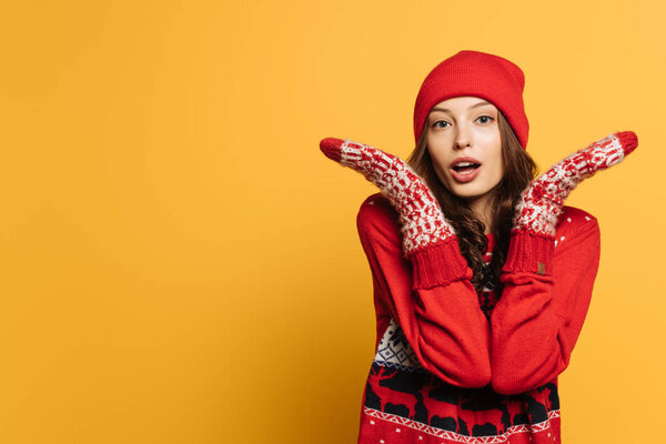 confused girl in red ornamental sweater and mittens showing shrug gesture on yellow background