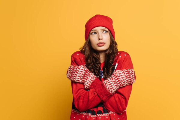 displeased girl in red ornamental sweater and mittens hugging herself while feeling cold isolated on yellow