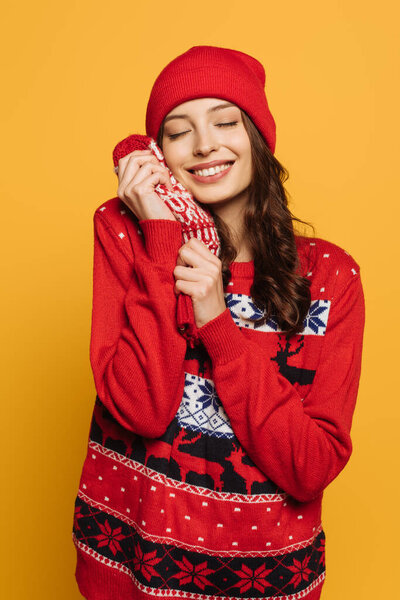 happy girl in hat and red ornamental sweater holding mittens near face with closed eyes isolated on yellow