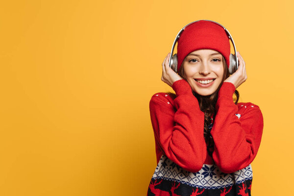 smiling girl in hat and red ornamental sweater listening music in wireless headphones on yellow background